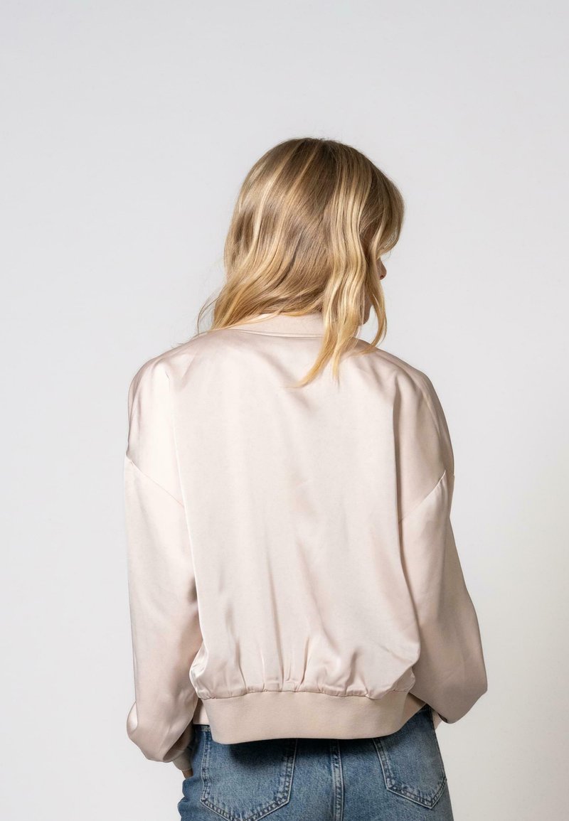 Giacca bomber beige chiaro in satin morbido, con spalle cadenti, orlo a coste e maniche lunghe. Vista da dietro, indossata con jeans in denim.
