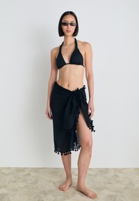 Svart bikini med halterneck-design, kombinert med en gjennomsiktig svart sarong med pompom-detaljer langs bunnen, stående på en steinlagt gulv.