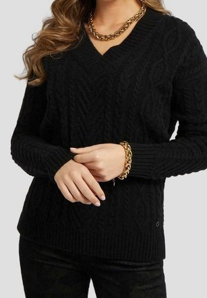 Femme portant un pull noir à col en V en maille torsadée avec un collier en chaîne dorée et un bracelet assorti, les mains délicatement croisées à la taille.