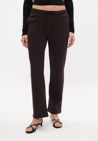 Brūnas sporta bikses ar auklu viduklī, taisna griezuma un mīksta tekstūra, kombinētas ar melnām sandālēm ar siksniņām.