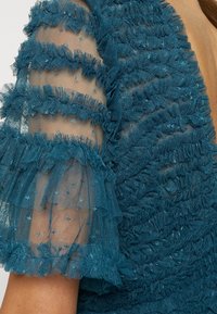 Mouw van een petrolblauwe jurk met doorschijnende stof en gerimpelde, getextureerde strepen die over de huid liggen.