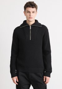 Suéter negro de canalé con un alto cuello y media cremallera. Cuenta con mangas largas y una textura gruesa de punto.