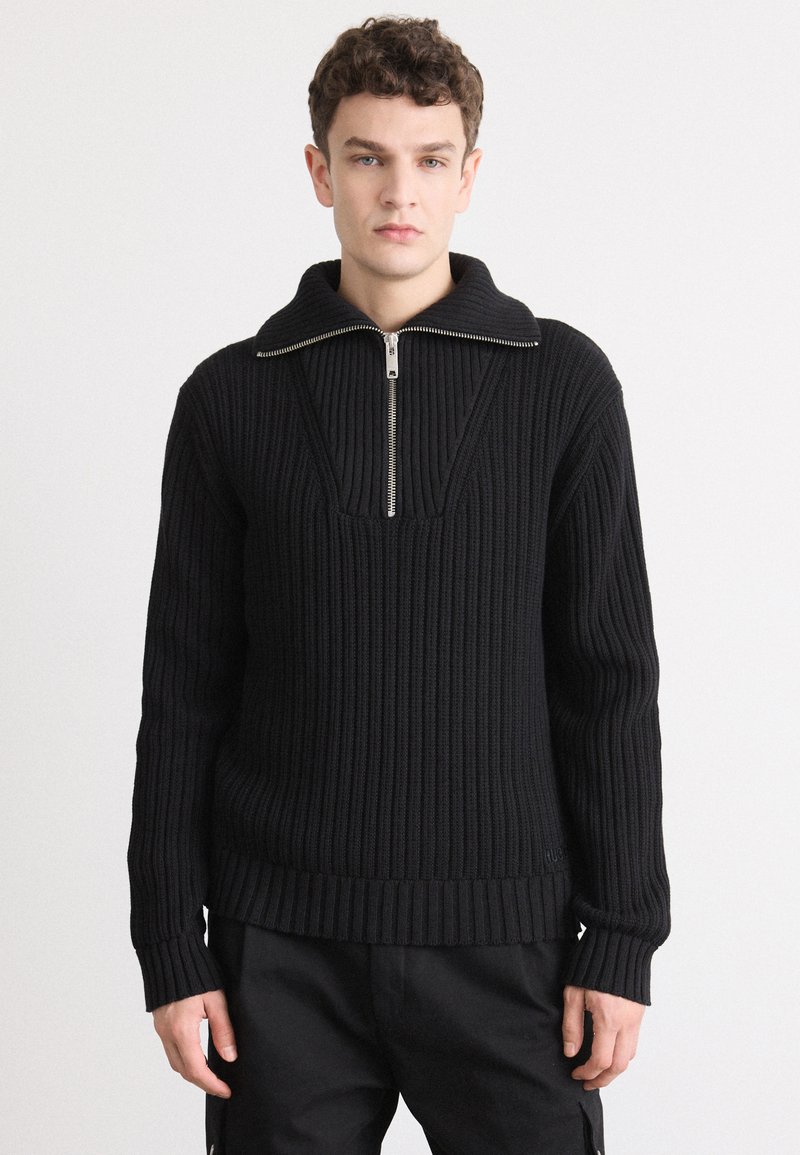Suéter negro de canalé con un alto cuello y media cremallera. Cuenta con mangas largas y una textura gruesa de punto.