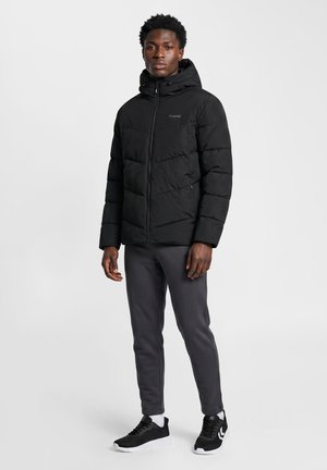 Mann steht, trägt eine schwarze Steppjacke mit Kapuze, dunkle Hosen, weiße Socken und schwarze Sneaker mit weißen Sohlen vor einem einfarbigen hellen Hintergrund.