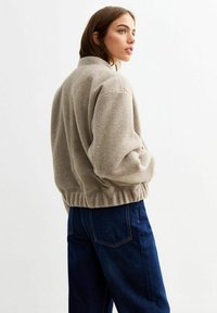 Beige zip-jacka i mjukt material, med en figurnära nederkant, sänkta axlar och ribbstickade manschetter, i kombination med mörkblå vida jeans.
