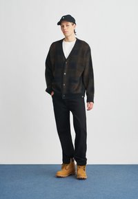 Svart og brunt mønstrete cardigan med V-hals, knappeåpning og lommer, kombinert med svarte bukser og brune snøresko.