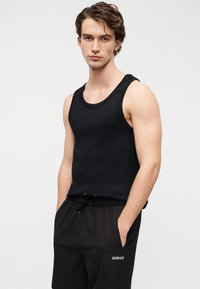 Μαύρο τοπ χωρίς μανίκια με ριμπ υφή, συνδυασμένο με μαύρο σορτς με κορδόνι, που φέρει λευκό λογότυπο. Το μοντέλο στέκεται σε ουδέτερο φόντο.