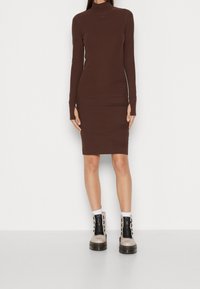 Figurformad brun klänning med långa ärmar och hög krage, tillverkad av slät tyg. Buren med kraftiga svarta och beige stövlar.