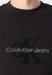 Czarna bluza z ribbingu przy dekolcie. Posiada haftowane logo: "Calvin Klein Jeans" w białym kolorze oraz "K" w strukturalnej czarnej nitce.