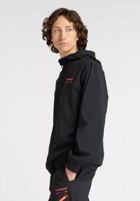 Schwarze leichte Jacke mit Kapuze, elastischen Bündchen und einem roten Logo auf der Brust. Das Material scheint glatt und wasserabweisend zu sein.