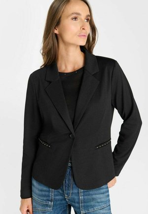 Vrouw met lichtbruin haar die een aangesloten zwarte blazer draagt over een zwart topje en blauwe spijkerbroek, staand met één hand in de zij, kijkend naar de zijkant.