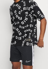 Czarna koszulka z białym napisem "PARIS" i rozsianymi logo Jumpman, zestawiona z czarnymi szortami Nike z białymi paskami po bokach i logo.
