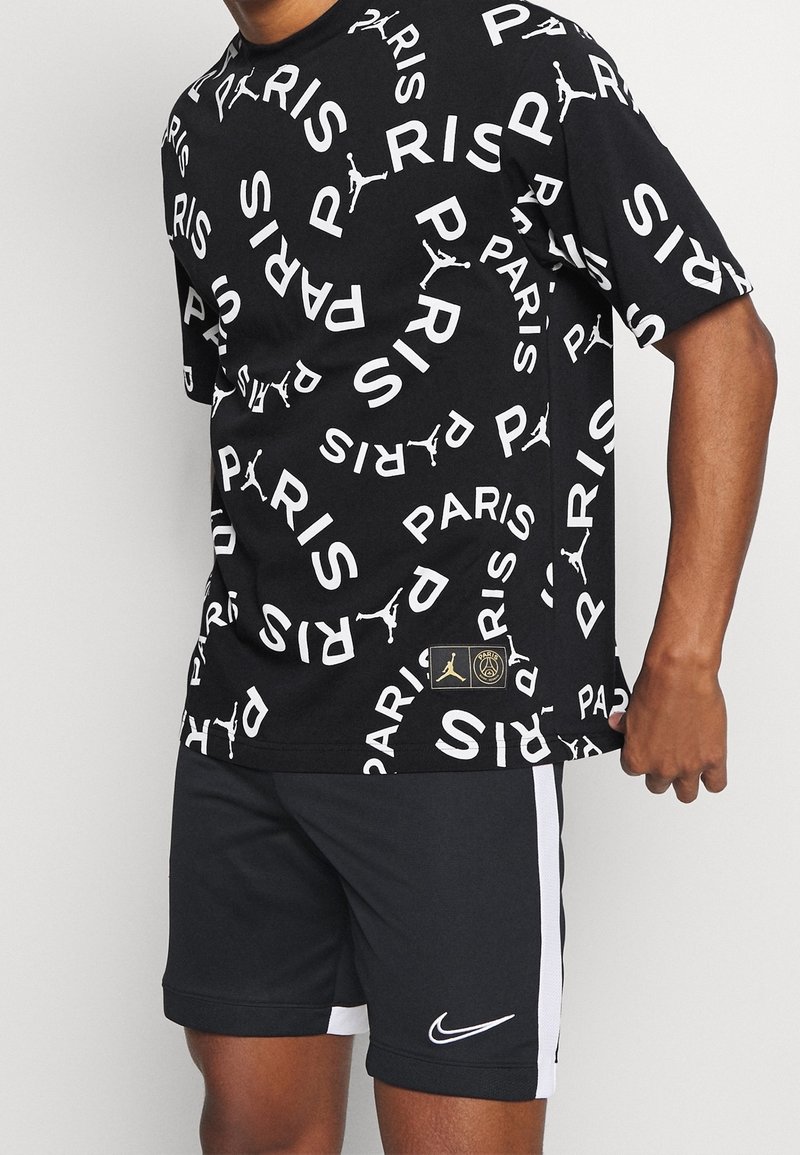 Czarna koszulka z białym napisem "PARIS" i rozsianymi logo Jumpman, zestawiona z czarnymi szortami Nike z białymi paskami po bokach i logo.