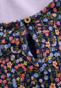 Prenda azul marino con estampado floral en rosa, amarillo y azul que presenta un pequeño botón de madera en un fondo de tela de lavanda acanalada.