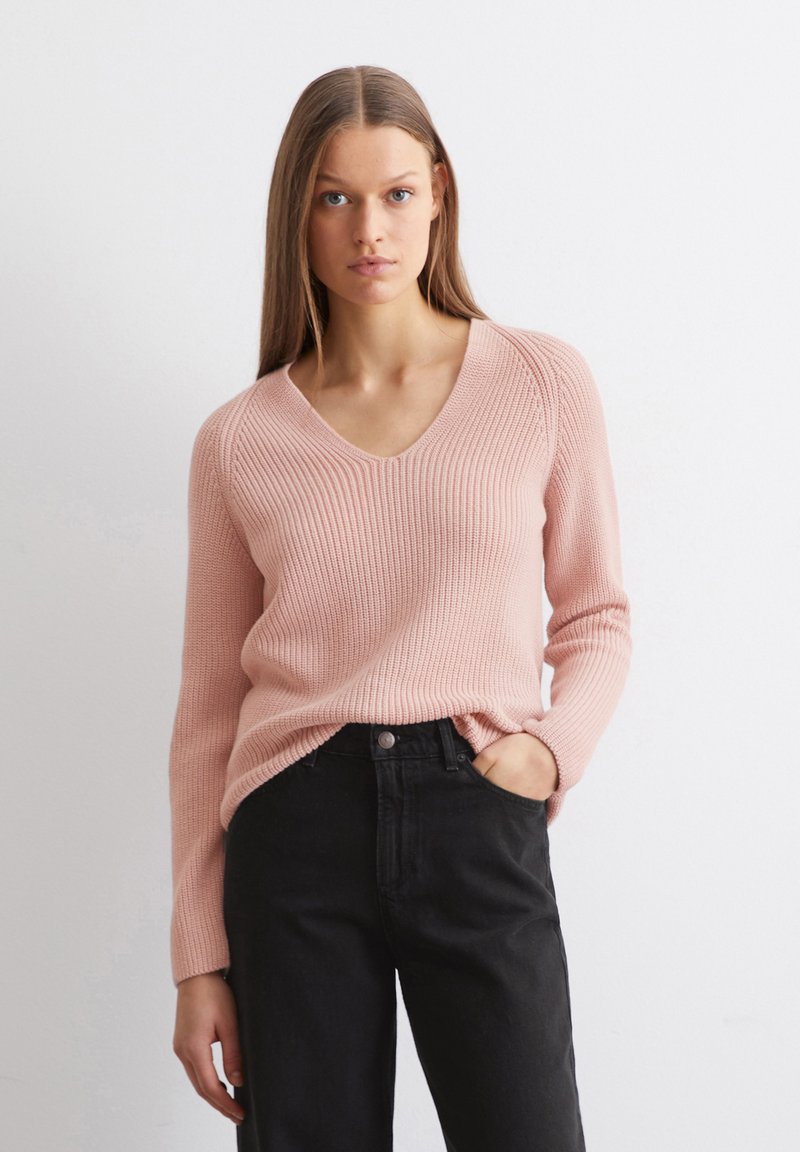 Hellrosa gestrickter Pullover mit V-Ausschnitt und geripptem Design, kombiniert mit schwarzen hochgeschnittenen Jeans. Langer Ärmel und lockere Passform.