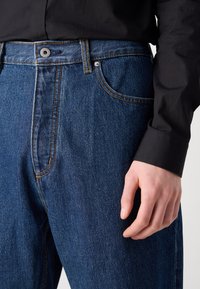 Blå denim jeans har en lige pasform, synlig syning i en kontrastfarve og en knaplukning i taljen med metal detaljer.