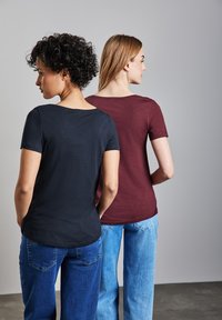 Due donne stanno schiena contro schiena, indossando magliette a maniche corte; una è vestita di nero e l'altra di bordeaux, abbinate a jeans blu.
