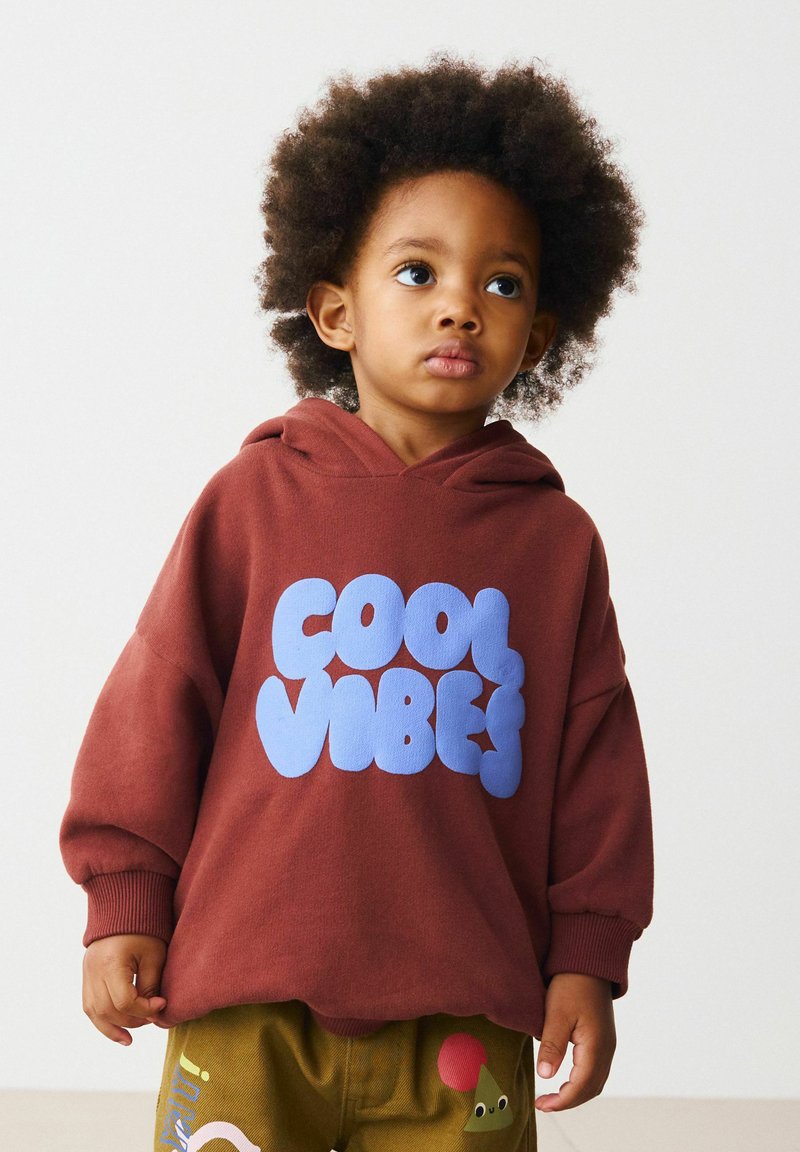 Haut à capuche marron avec le texte bleu "COOL VIBES", manches longues et coupe décontractée. Assorti à un pantalon vert olive avec des accents ludiques.
