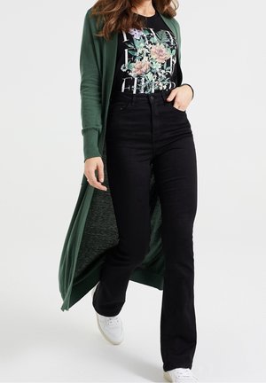 Femme portant un long cardigan vert, un t-shirt noir à motif floral, un jean noir taille haute et des baskets blanches marchant vers l'avant.