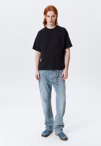 Zwarte t-shirt met korte mouwen gecombineerd met lichtblauwe denim jeans met een metalen sleutelhanger detail op de tailleband en blauwe klompen.