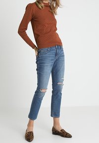 Pull marron texturé, jean déchiré bleu et ballerines marron à imprimé léopard. Concentration sur les détails des tissus et les éléments de design.