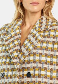 Spleciony płaszcz z wzorem w kratę w odcieniach brązu, żółci i bieli. Podwójne czarne guziki oraz fakturowany, wyraźny kołnierz.