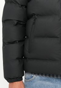 Veste matelassée noire avec un design matelassé, dotée d'une poche zippée et de poignets élastiques, associée à un jean en denim bleu.