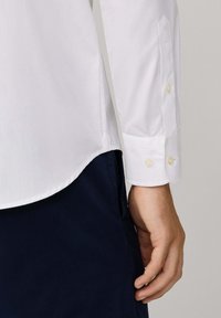 Camisa blanca de manga larga con puños abotonados, que presenta una textura suave y un dobladillo curvado. Usada sobre pantalones azul marino oscuros.