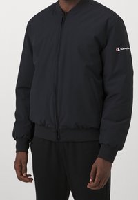 Svart bomberjacka i slät tyg, med en frontdragkedja, ribbade muddar och nederkant, samt en liten logotyp på vänster ärm.