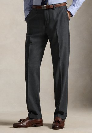 Bărbat purtând pantaloni eleganți gri închis, curea maro din piele, cămașă albastră cu dungi și mocasini maro cu ciucuri, mâinile în buzunare.