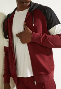 Sudadera con capucha granate con detalles en negro y blanco. Cuenta con capucha ajustable con cordón, puños acanalados y un pequeño logo en el pecho.