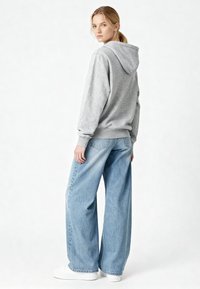 Felpa grigia in cotone con cappuccio regolabile, abbinata a jeans larghi di colore blu chiaro e sneakers bianche. Design semplice e vestibilità casual.