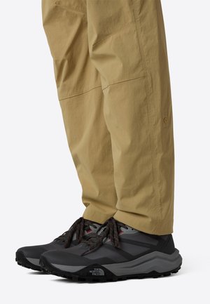 Osoba v světle hnědých kalhotách a černo-šedých turistických botách The North Face s červenými šňůrkami stojí na bílém pozadí.