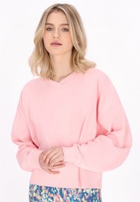Felpa oversize rosa con scollo a V, maniche larghe e polsini a costine, abbinata a un pantalone con fantasia multicolore. Tessuto morbido.