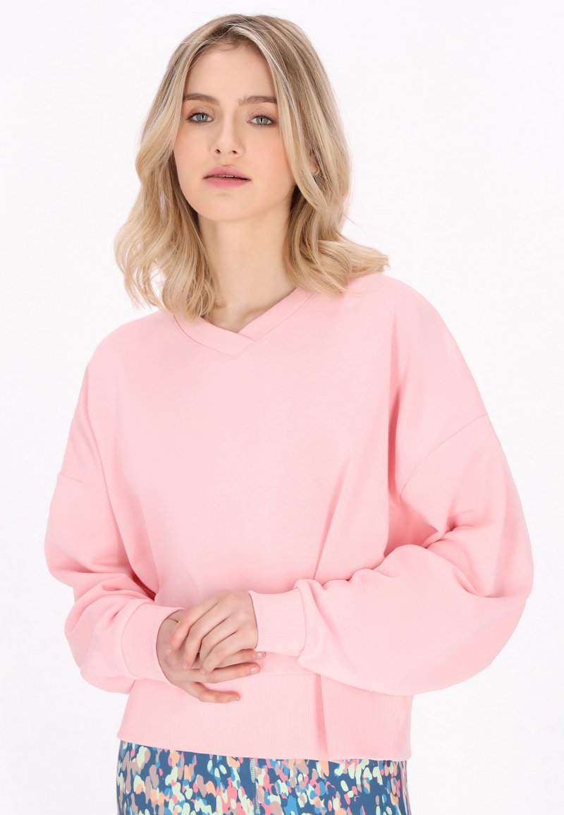 Felpa oversize rosa con scollo a V, maniche larghe e polsini a costine, abbinata a un pantalone con fantasia multicolore. Tessuto morbido.