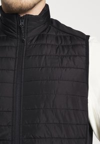 Schwarze gepolsterte Weste mit hohem Kragen, ausgestattet mit einem durchgehenden Reißverschluss und horizontalem Steppmuster. Enthält ein dezentes Logodetail auf der Brust.