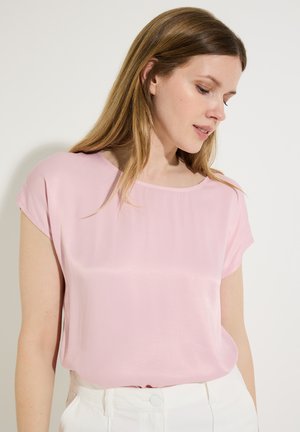 Frau mit glattem, hellbraunem Haar, die eine hellrosa kurzärmelige Bluse und weiße Hose trägt und auf einen weißen Hintergrund nach unten schaut.