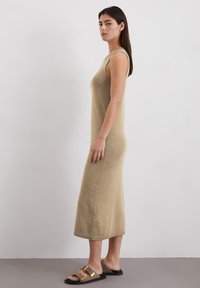 Robe en tricot beige, sans manches, avec une silhouette cintrée et un ourlet droit. Les sandales présentent une sangle marron et des accents de boucle en or.