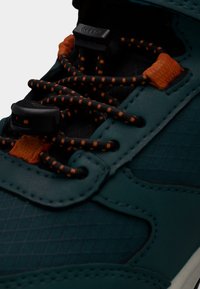 Αθλητικό παπούτσι σε τιρκουάζ χρώμα με υφή από συνθετικό υλικό, μαύρες λεπτομέρειες και πορτοκαλί κορδόνια με κουμπί ρύθμισης για ασφαλές δέσιμο.