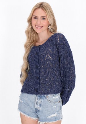 Marineblauer Strickcardigan mit einem Spitzenmuster, Rundhalsausschnitt und vier vorderen Knöpfen. Kombiniert mit zerfetzten Denim-Shorts.