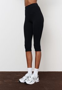 Fekete, térd alatt érő leggings, rugalmas anyagból, sima textúrával és magas derékkal. Fehér sportcipővel és zoknival viselve.