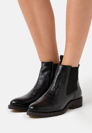 Černé kotníkové boty z texturované kůže s elastickými bočními panely, nošené na bosých nohách, s nízkými podpatky a podrážkami zdobenými nýty.