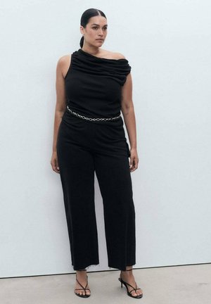 Black off-the-shoulder top with draped fabric, paired with wide-leg black trousers and a metallic chain belt, worn with thin-strap sandals.