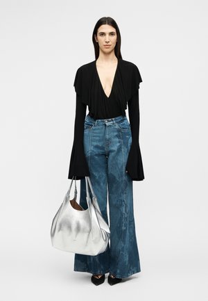 Ženska, oblečena v črno majico z globokim izrezom v obliki črke V, širokimi zvončastimi rokavi, modre kavbojke z visokim pasom in širokimi hlačnicami, črne konične čevlje, nosi veliko srebrno torbico.