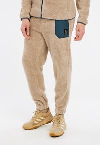 Pantalones de forro polar beige con pierna ajustada, con un bolsillo lateral de color azul oscuro. La tela tiene una apariencia texturizada y esponjosa.