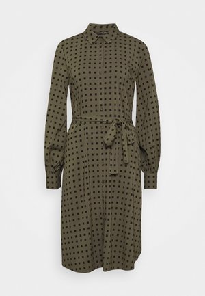Robe-chemise vert olive longueur genou avec motif carré noir, manches longues, boutonnée devant, col et ceinture nouée à la taille.