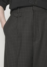 Gros plan sur un pantalon noir à carreaux avec de petites lignes blanches formant une grille, avec une poche latérale boutonnée et une main glissée dans une autre poche.