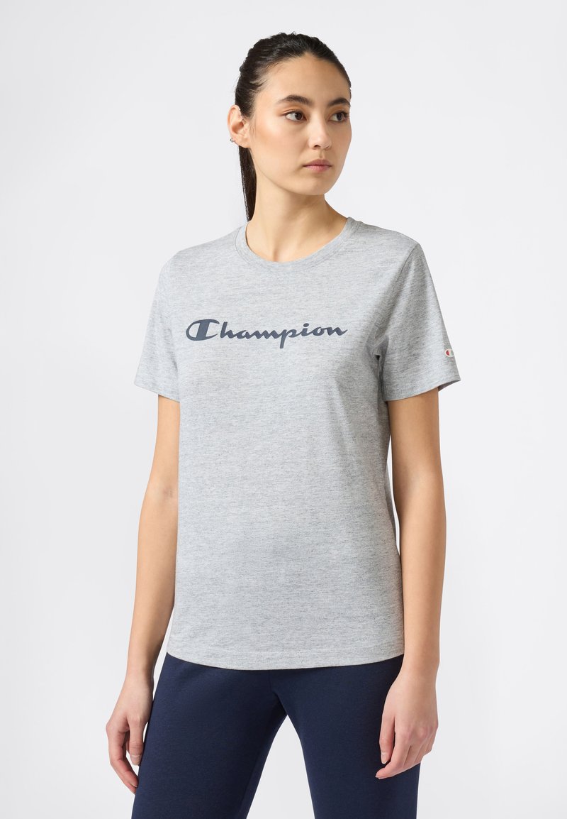 Młoda kobieta z ciemnymi włosami związanymi w kucyk, ubrana w jasnoszary t-shirt z logo Champion i ciemne spodnie, stoi na tle białym, patrząc w prawo.