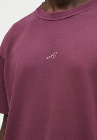 Κοντινή λήψη ενός ατόμου που φοράει απλό μαρόν μπλουζάκι με ένα μικρό, μινιμαλιστικό λευκό γεωμετρικό λογότυπο στο στήθος.