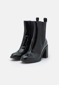 Black patent leather ankle boots with a high block heel, elastic side panels, and a logo detail on the outer side. Smooth, shiny texture.